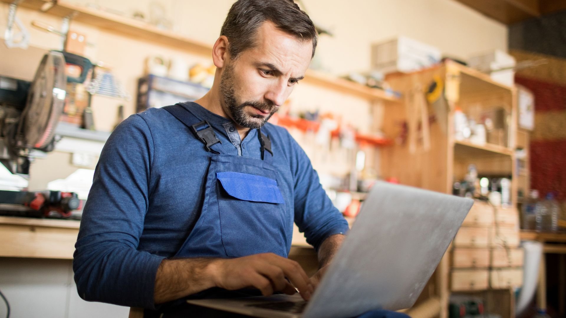 Worker in blue uniform using laptop in workshop with tools around
