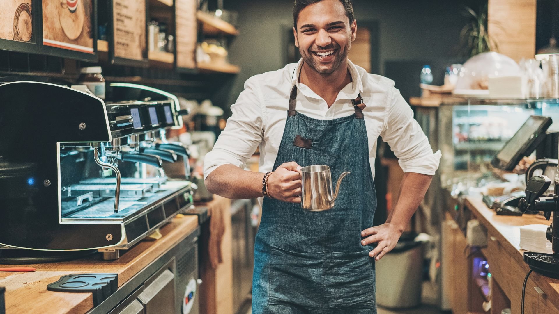 Smiling barista holding milk pitcher near espresso machine in cafe
